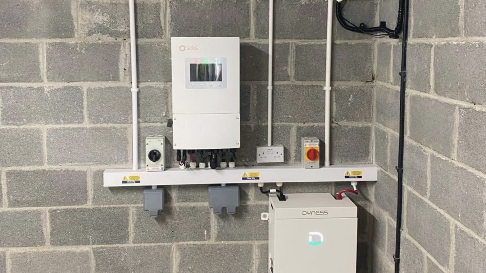 A white Solis inverter on a block wall above white trunking with isolator switches, and a Dyness battery on the floor below
